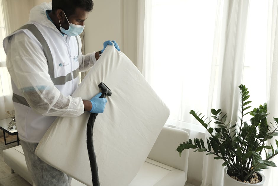 A professional cleaner wearing a face mask, gloves, and a white protective suit is performing surface cleaning on a mattress using a handheld vacuum or steam cleaning device. The setting is a bright,