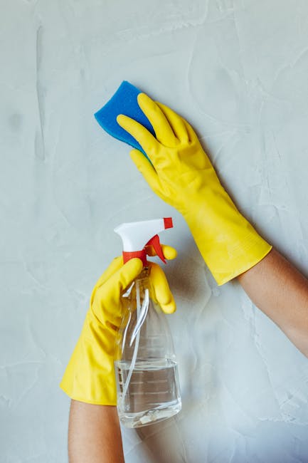 A professional cleaner wearing a face mask, gloves, and a white protective suit is performing surface cleaning on a mattress using a handheld vacuum or steam cleaning device. The setting is a bright,